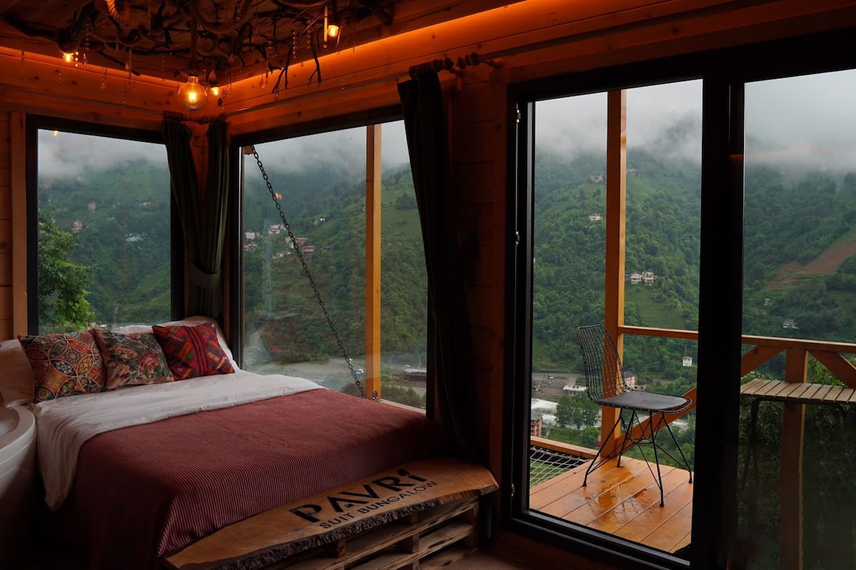 A bedroom with a large bed, a bench with 'PAVRI SUIT BUNGALON' text, and a view of a green landscape through large windows.