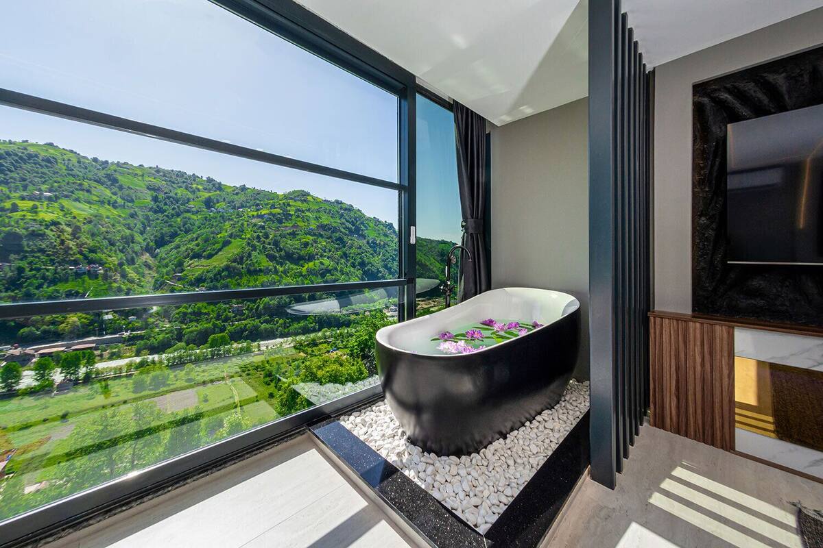 A modern bathroom with a freestanding bathtub, large windows, and a view of a green landscape.