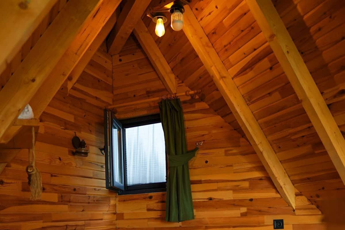 A wooden cabin bedroom with a bed, a window with curtains, and a hanging light fixture.