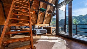 A cozy cabin interior with a wooden staircase, a gray sofa, and a large window overlooking a scenic landscape. - Pavri Suit Bungalov (Ardesen)