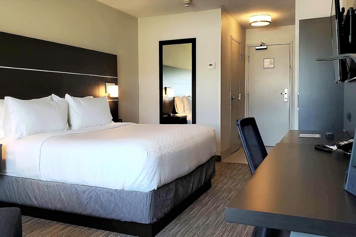 A hotel room with a large bed, a desk, a chair, a TV, and a mirror.