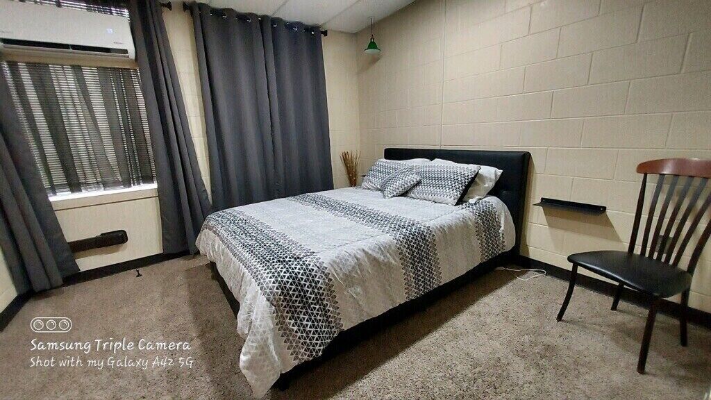 A bedroom with a bed, a chair, a window with curtains, and an air conditioning unit.