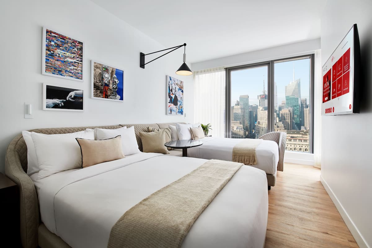 Ein modernes Hotelzimmer mit einem großen Bett, einem Einzelbett, einem runden Tisch und Blick auf die Stadt durch große Fenster.