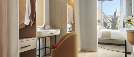 A modern hotel room with a city view, a vanity with a mirror, a desk, and a comfortable seating area.