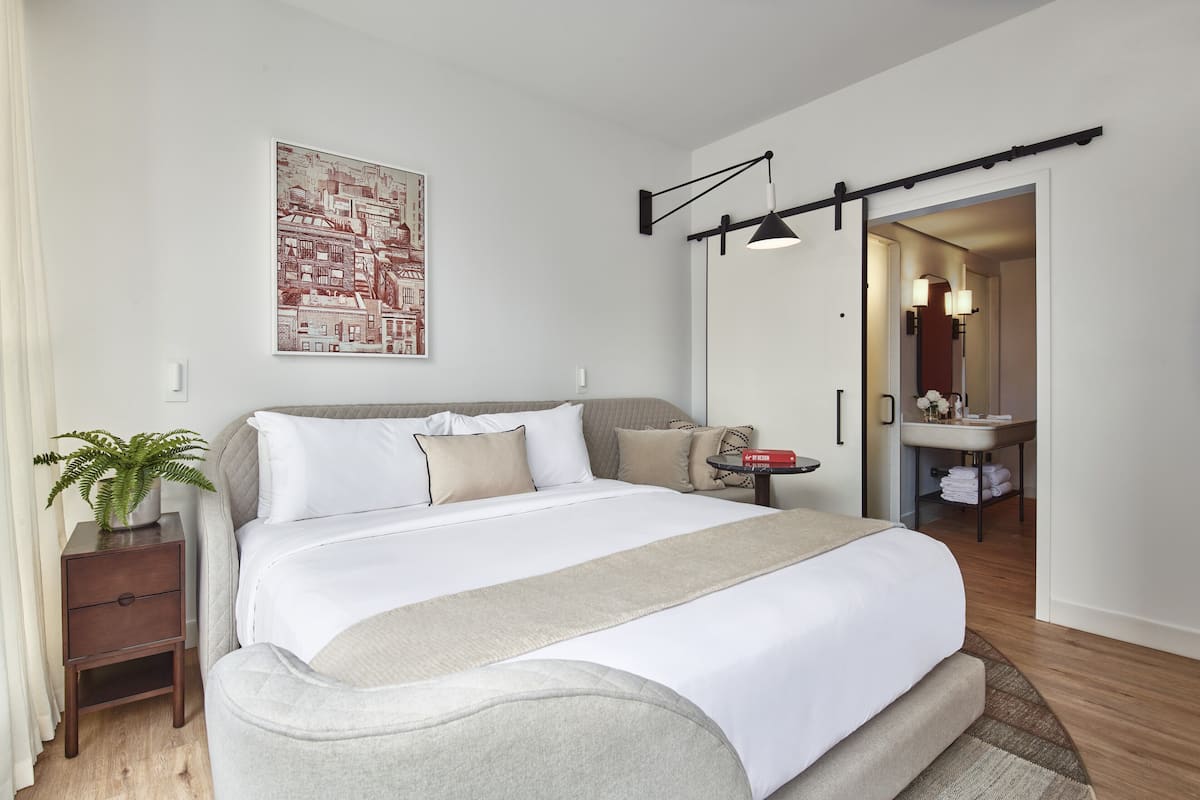 Ein modernes Hotelzimmer mit einem großen Bett, einem Nachttisch mit Pflanze, einem Wandbild und einem durchgegangene Tür erkennbaren Badezimmer.