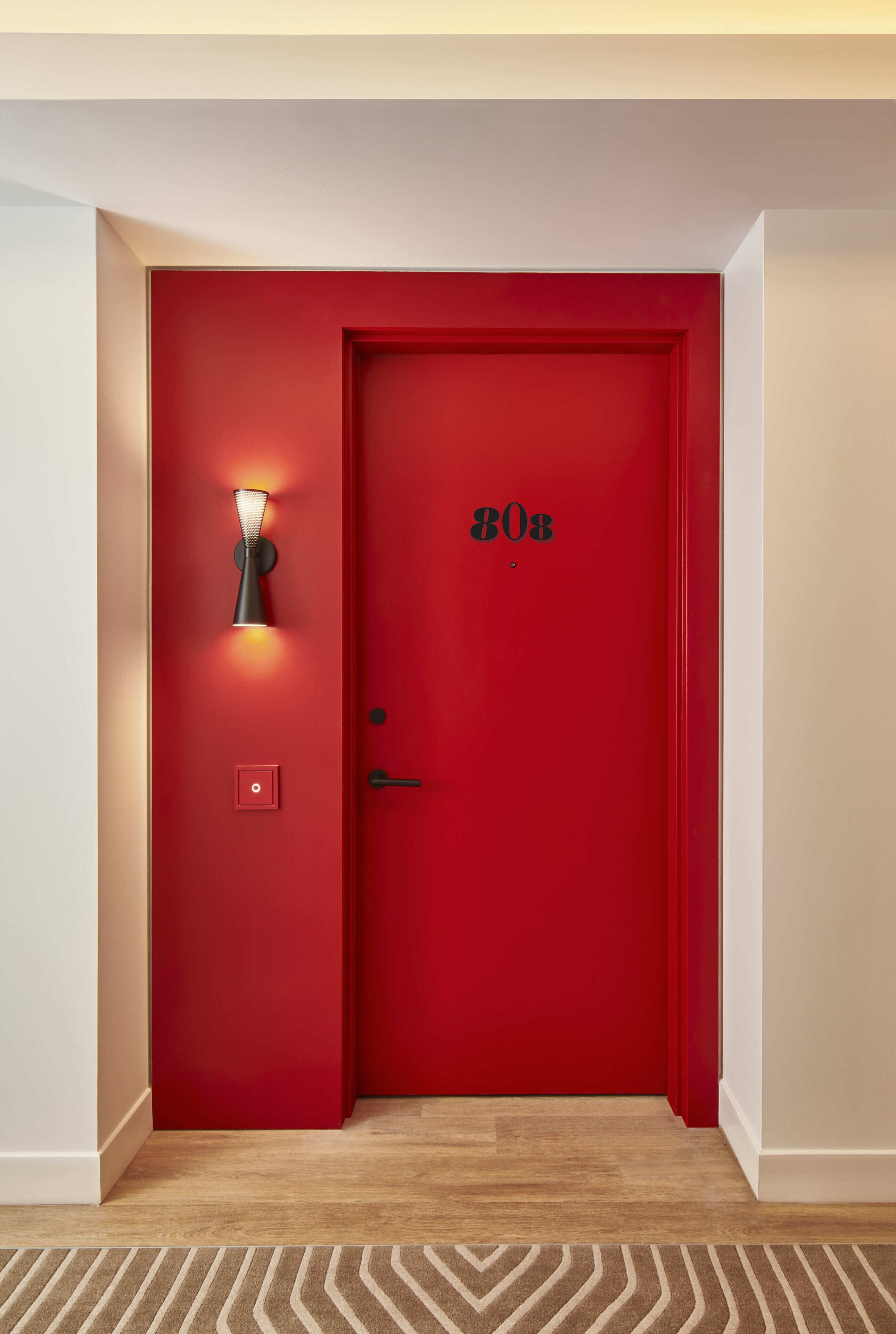 A red door with a number plate, a wall-mounted light fixture, and a power outlet.