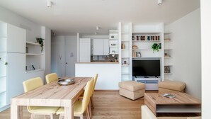 Een moderne woonruimte met een houten eettafel, gele stoelen, een beige bank, een televisie en ingebouwde planken met boeken en planten.