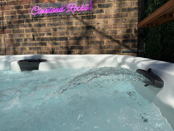 A hot tub with water flowing out, set against a brick wall with a neon sign that reads "Clandestine Rocks!".