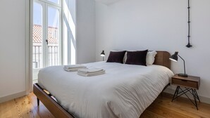 Et soveværelse med en stor seng, to sengeborde, et natbord i træ og udsigt til en bygning gennem et vindue.