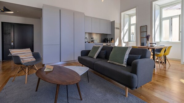 A modern living room with a grey sofa, a wooden coffee table, and a black chair. There is a kitchen area with white cabinets and a dining table with yellow chairs.