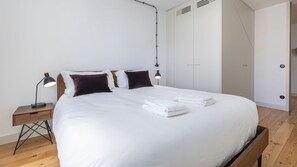 A neatly made bed with white linens, two dark pillows, and a stack of folded white towels. There is a wooden nightstand with a black lamp to the left.