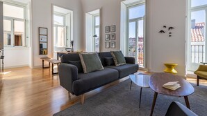En moderne stue med en mørkegrå sofa, et rundt sofabord og en træbænk. Store vinduer giver naturligt lys og udsigt til bygninger.