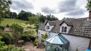 A white house with a stone patio, a glass greenhouse, and a view of a green hill. - Beautiful Cotswolds Cottage & Garden (Horsley)
