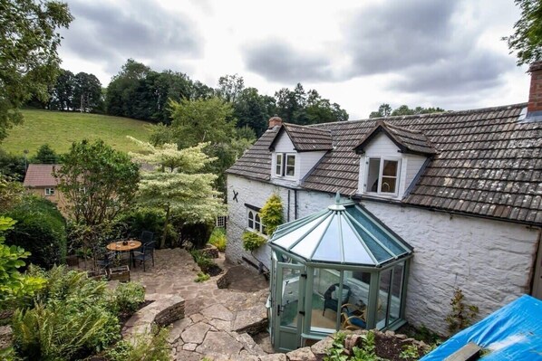 Une maison blanche avec un patio en pierre, une serre en verre et une vue sur une colline verdoyante.
