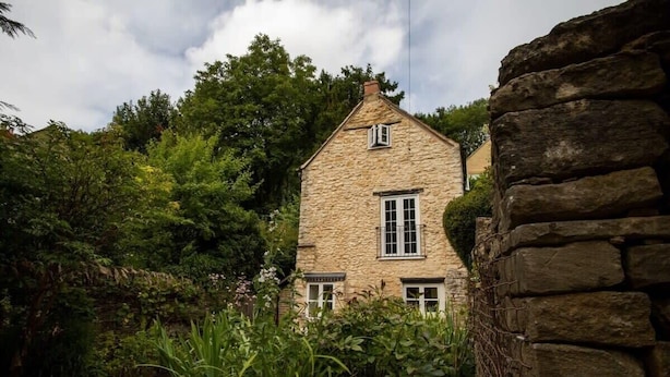 A stone house with a gabled roof, surrounded by lush greenery and a stone wall. - Beautiful Cotswolds Cottage & Garden (Horsley)