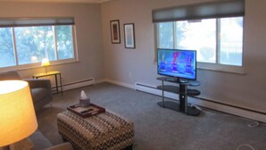Een woonkamer met een televisie, een lamp, een gestreept voetenbankje en een klein tafeltje.