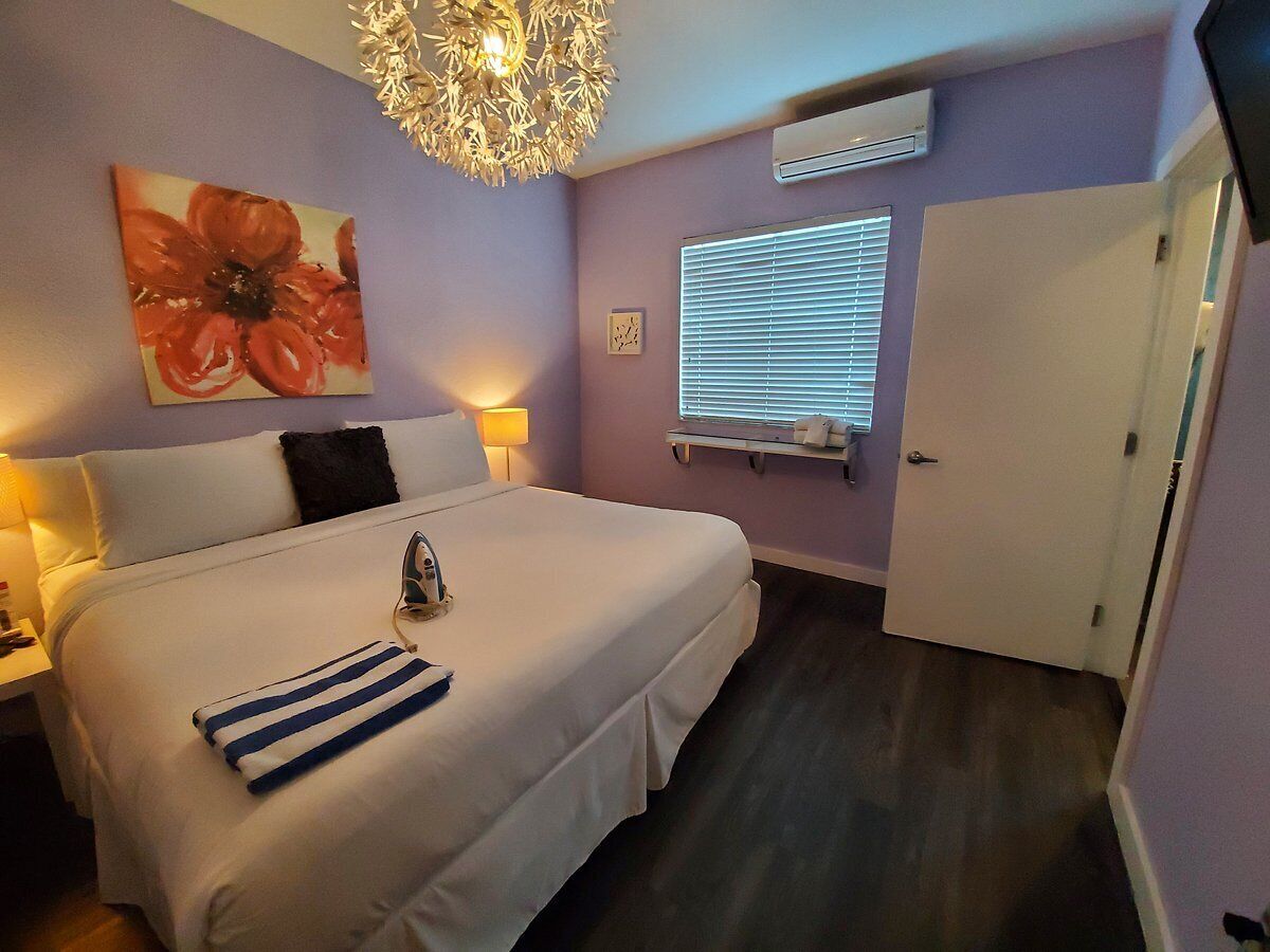 A bedroom with a bed, bedside table, lamp, artwork, window, and door.