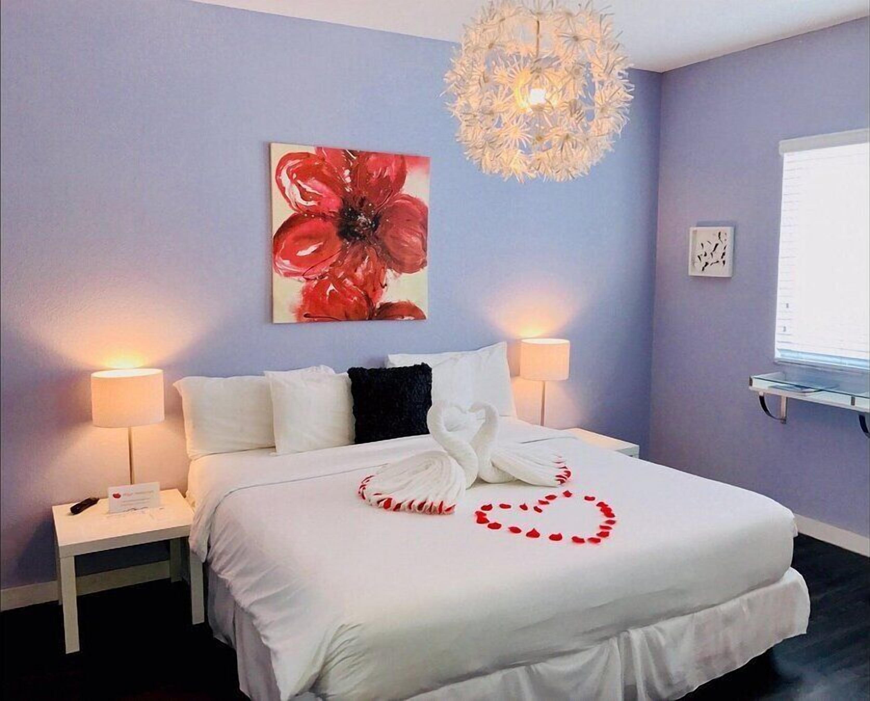 Een hotelkamer met een bed, twee bedlampen, een kroonluchter, een schilderij van een rode bloem en een hartvormige decoratie op het bed.