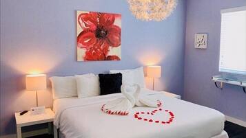 Een hotelkamer met een bed, twee bedlampen, een kroonluchter, een schilderij van een rode bloem en een hartvormige decoratie op het bed.