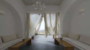A minimalist room with a large window, white curtains, a wooden bench, and a chandelier. - Mykonos Teatro Suites and Villa (Mykonos)