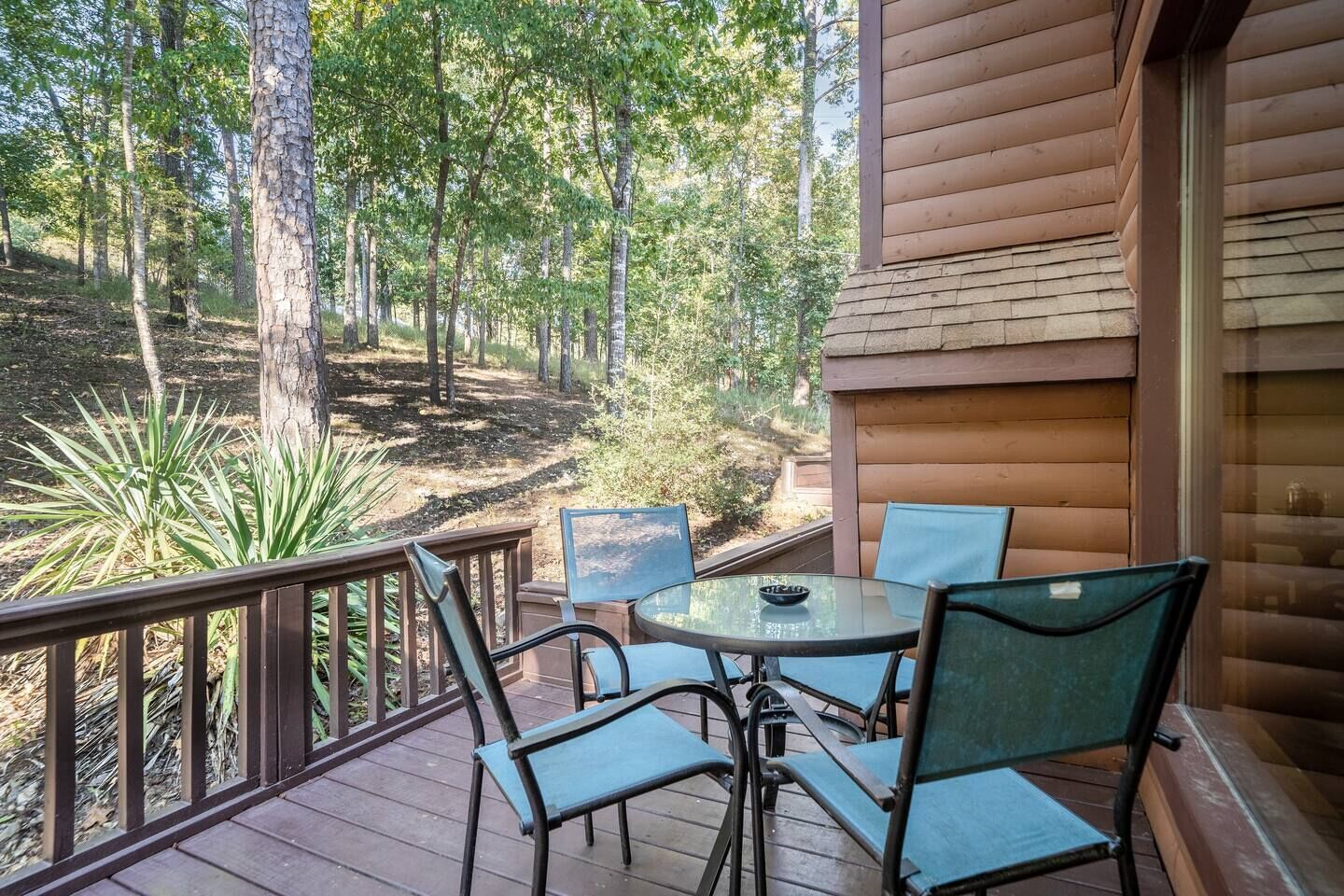Une terrasse en bois avec une table et des chaises en verre, donnant sur une zone boisée.