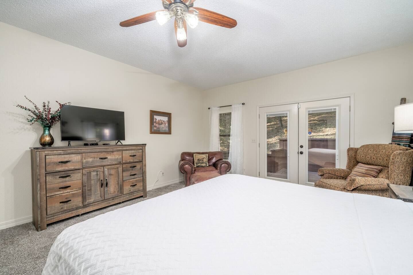 A bedroom with a large bed, a wooden dresser, a television, a leather armchair, and a ceiling fan.