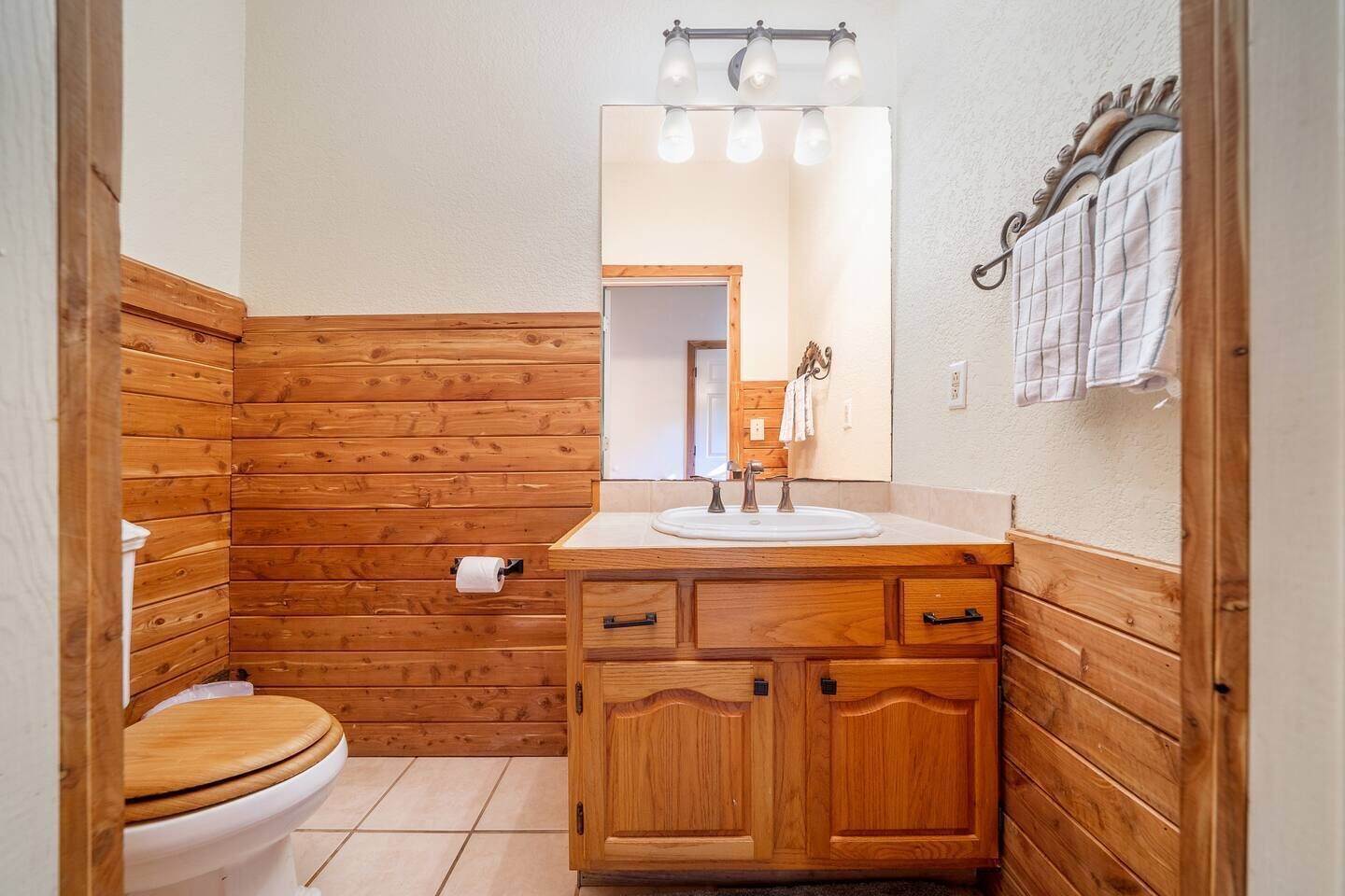 Een badkamer met houten lambrisering, een wit wastafelmeubel met twee kraan en een spiegel erboven, en een toilet met houten zitje.
