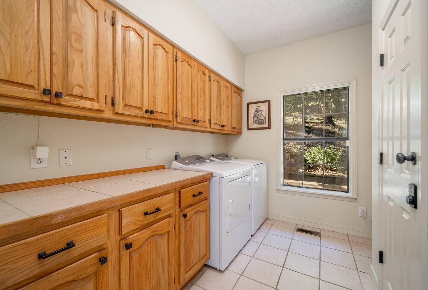 A kitchen with wooden cabinets, a washing machine, a countertop, and a window with a view of greenery.