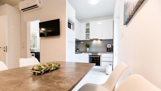 A modern kitchen with white cabinets, a built-in oven, and a flat-screen TV mounted above the counter. - L&L Apartments (Split)