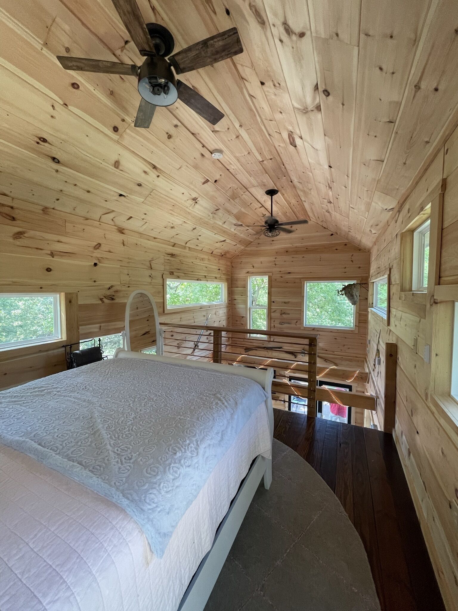 A wooden cabin bedroom with a bed, two windows, a ceiling fan, and a balcony.