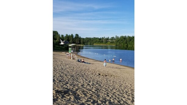 A sandy beach with a calm lake, a covered seating area, and a few people enjoying the water. - The Rameau 1er (Montréal)