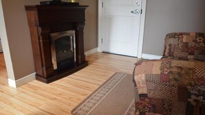 A room with a wooden floor, a fireplace, a door, and a patterned sofa. - Le Citadin (Rouyn-Noranda)