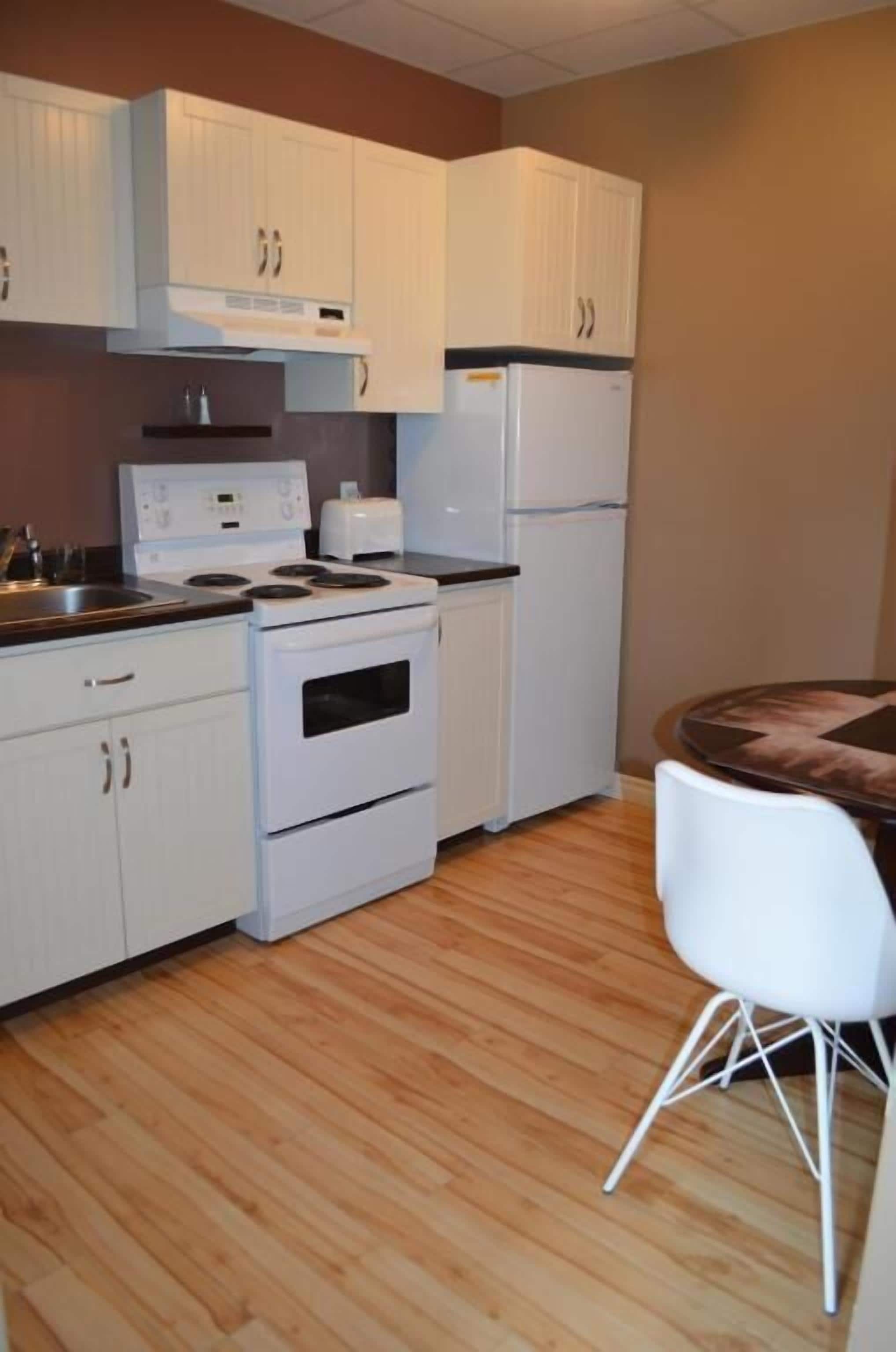 A compact kitchen with white cabinets, a stove, a refrigerator, and a dining area with a white chair.
