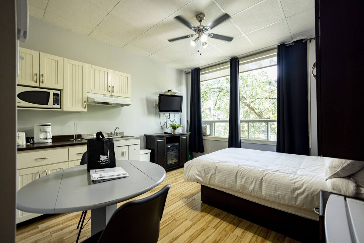 Habitación de hotel compacta con cama, un pequeño escritorio con silla, microondas, televisor y ventilador de techo.