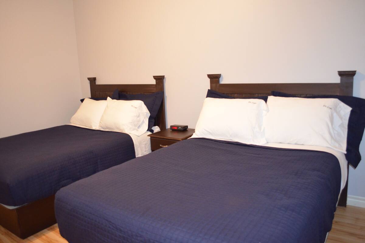 Dos camas individuales con ropa de cama azul oscuro, cabeceros de madera y una mesita de noche con un reloj entre ellas.