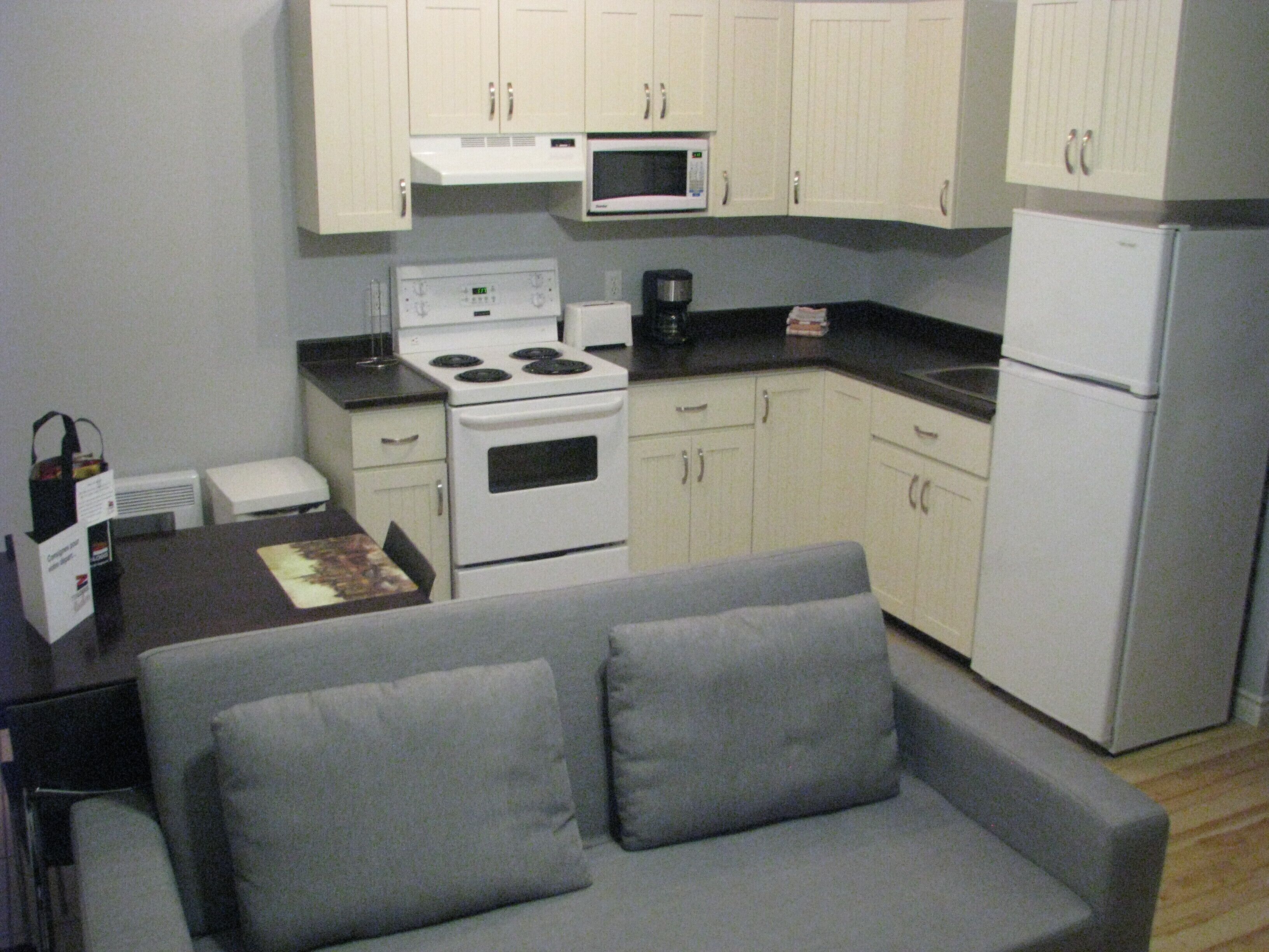 A compact kitchen with white cabinets, a microwave, a stove, and a refrigerator. There is a gray sofa in the living area.
