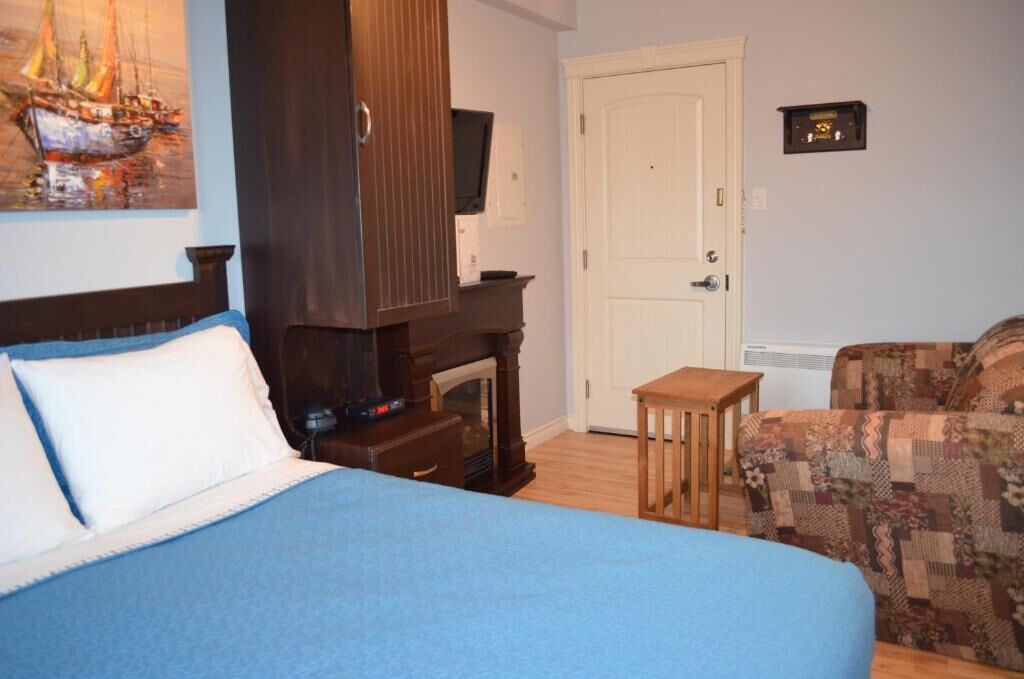 A bedroom with a bed, a small wooden table, a patterned armchair, a fireplace, a door, and a wall-mounted television.