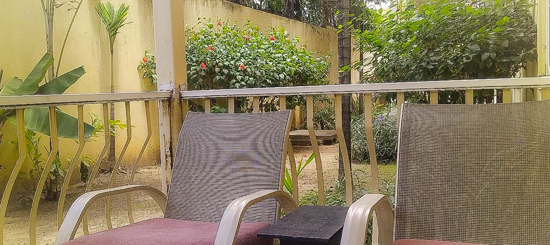 Un balcón con dos sillas de mimbre y una mesa, rodeado de plantas y una pared amarilla.