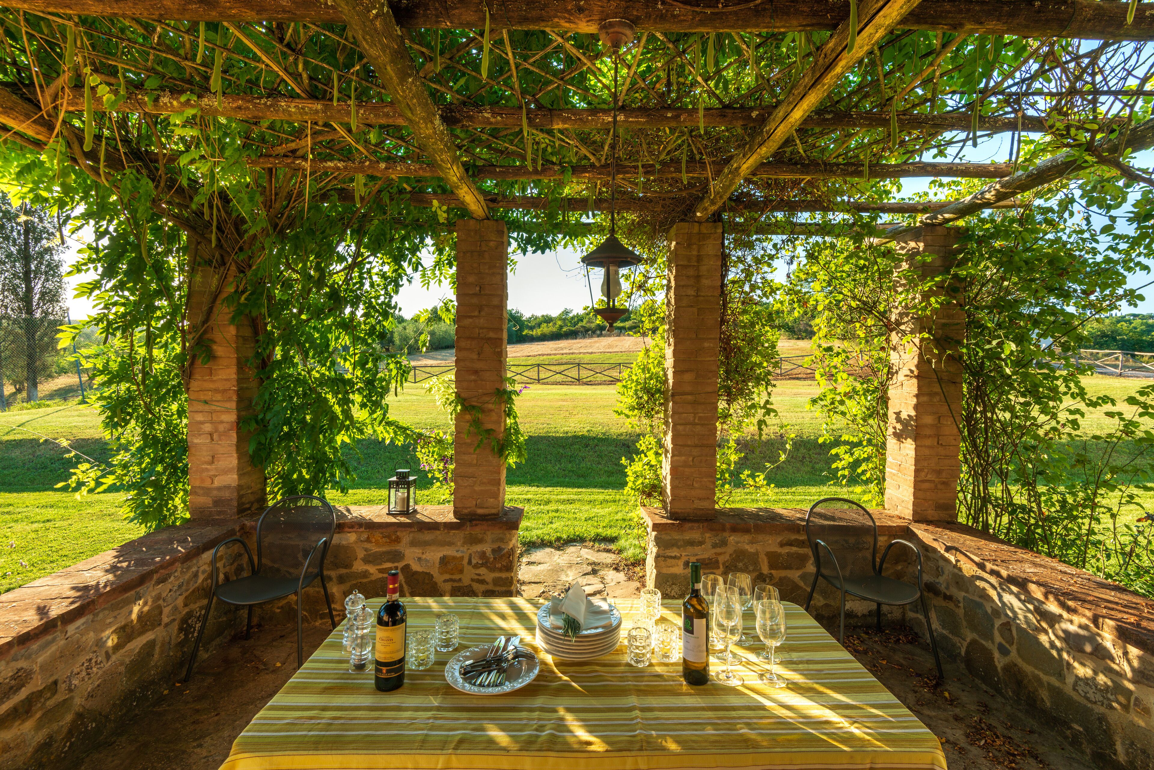 An outdoor dining area with a table set for two, surrounded by stone pillars and covered by a pergola.