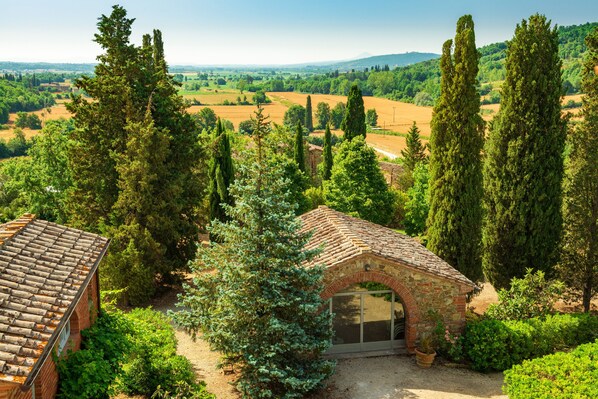 A rustic villa with a brick roof, surrounded by lush greenery and tall cypress trees.