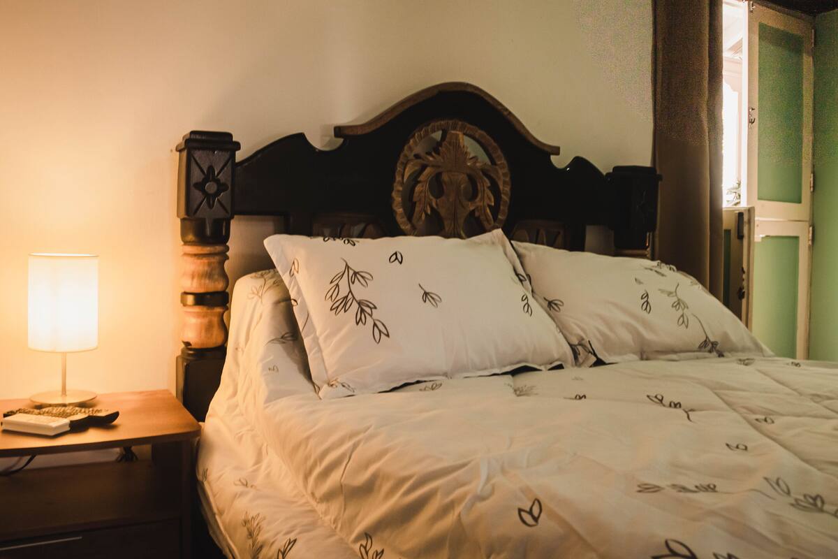 Una cama bien hecha con estampado floral, cabecera de madera, lámpara de noche y mesita de noche con un control remoto.