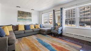 A modern living room with a sectional sofa, a patterned rug, and large windows offering a view of the city.
