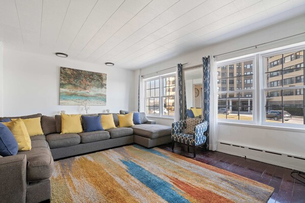 A modern living room with a sectional sofa, a patterned rug, and large windows offering a view of the city.
