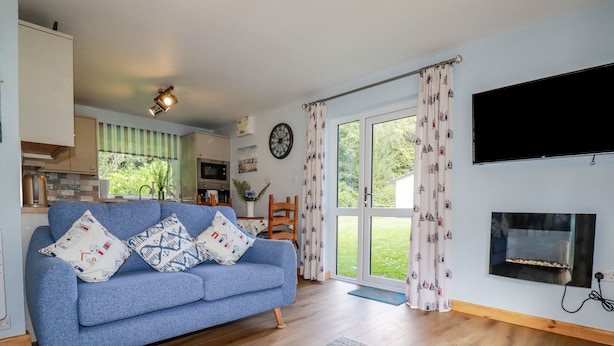 A modern living room with a blue sofa, wooden flooring, a fireplace, and a large window with curtains. - Woodside (Liskeard)