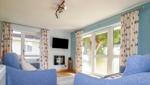 A living room with two blue armchairs, a flat-screen TV mounted on the wall, and a wooden cabinet. - Woodside (Liskeard)
