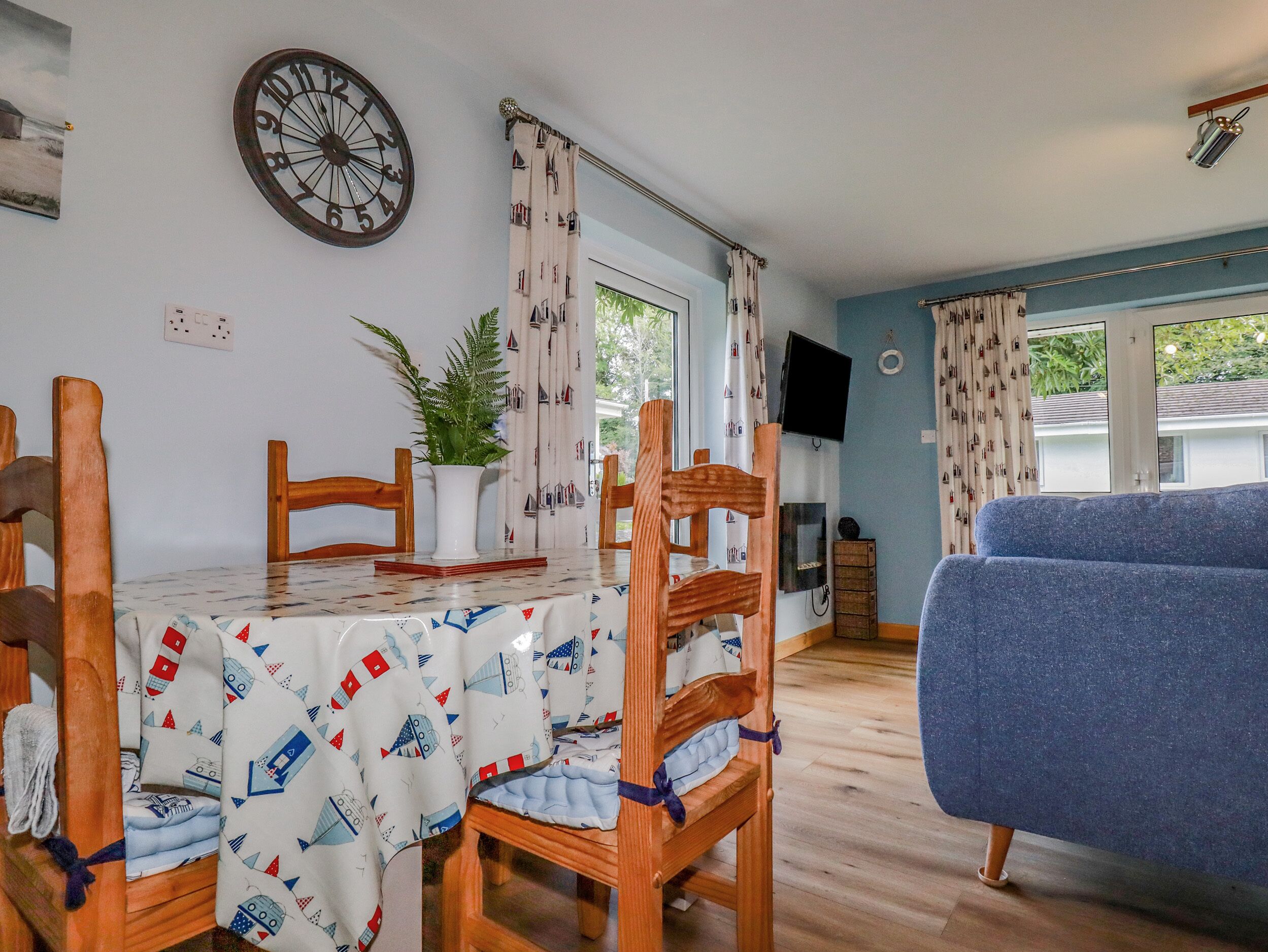 A dining area with a table covered by a nautical-themed tablecloth, wooden chairs, and a potted plant. - Woodside (Liskeard)