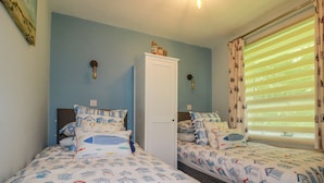 A bedroom with two beds, each with matching bedding featuring a nautical theme. There is a wardrobe and a window with blinds. - Woodside (Liskeard)