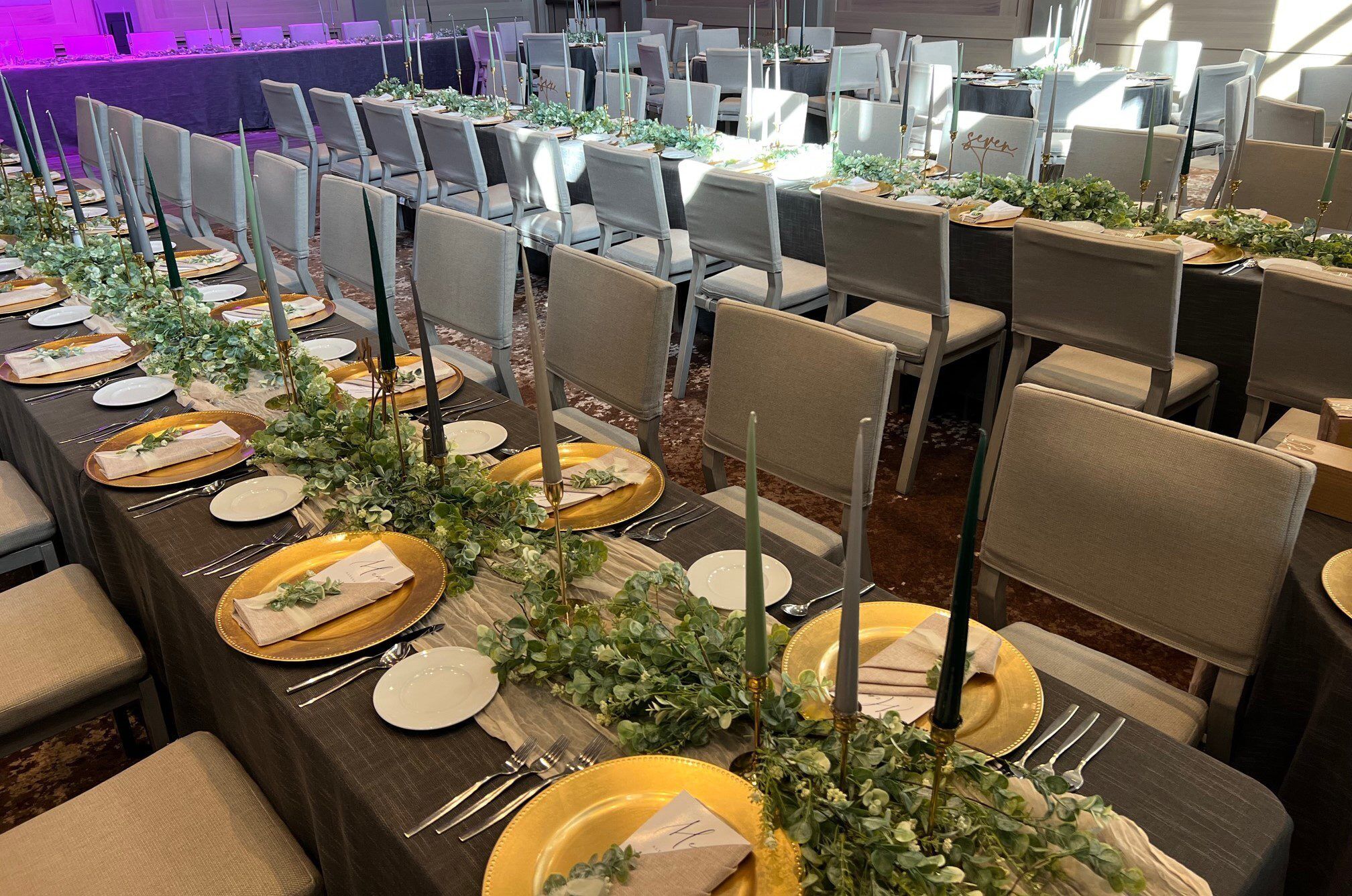 En festlokal med rader av bord uppdukade för ett formellt evenemang, med guldkantade tallrikar, bestick och gröna, dekorativa blomsterarrangemang i mitten av varje bord.