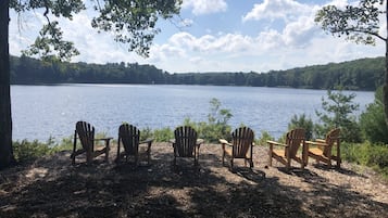 A lakeside view with wooden chairs facing the water, surrounded by trees and a clear sky.