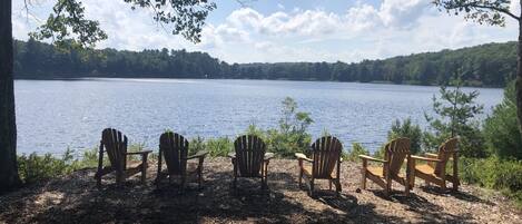 A lakeside view with wooden chairs facing the water, surrounded by trees and a clear sky.